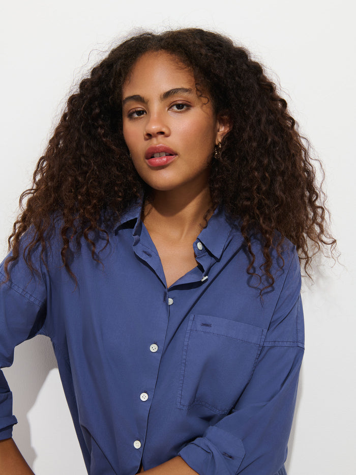 A woman with curly hair wears the Jo Shirt in Cotton Poplin, a blue 100% cotton button-up featuring French seams, as she looks at the camera against a plain white background.