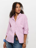 A woman with curly hair stands against a white background, wearing the Jo Shirt In Stripe Cotton Poplin—a loose-fitting, light pink striped 100% cotton button-up—paired with blue jeans. She looks to the side with one hand in her jeans pocket and a blue scrunchie on her wrist.