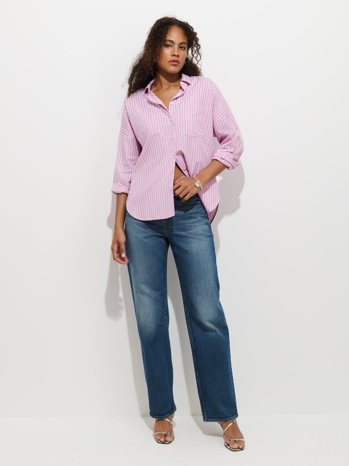 A person wearing the Jo Shirt In Stripe Cotton Poplin—a light pink striped 100% cotton button-up shirt with French seams—paired with blue jeans and sandals, stands against a plain white background, looking at the camera with a relaxed posture.