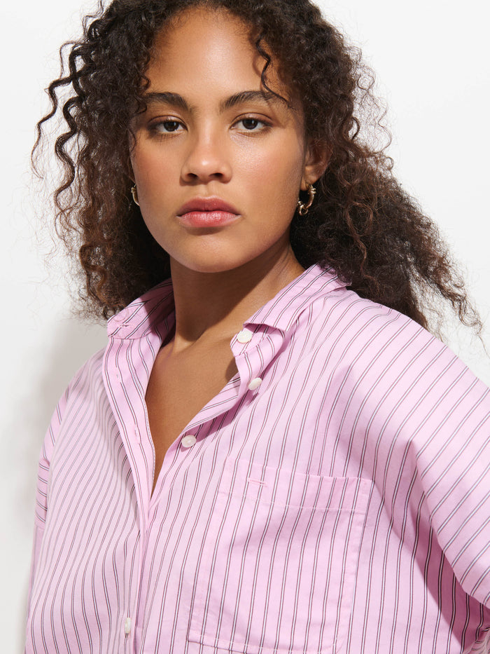 A woman with curly hair wears the Jo Shirt in Stripe Cotton Poplin, a light pink, pinstriped button-up crafted from 100% cotton. She gazes confidently at the camera against a plain white background.