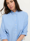 A woman with long brown hair wears the Jo Striped Shirt in Cotton Poplin—an oversized light blue shirt with white and red stripes—standing against a plain white background and looking at the camera.