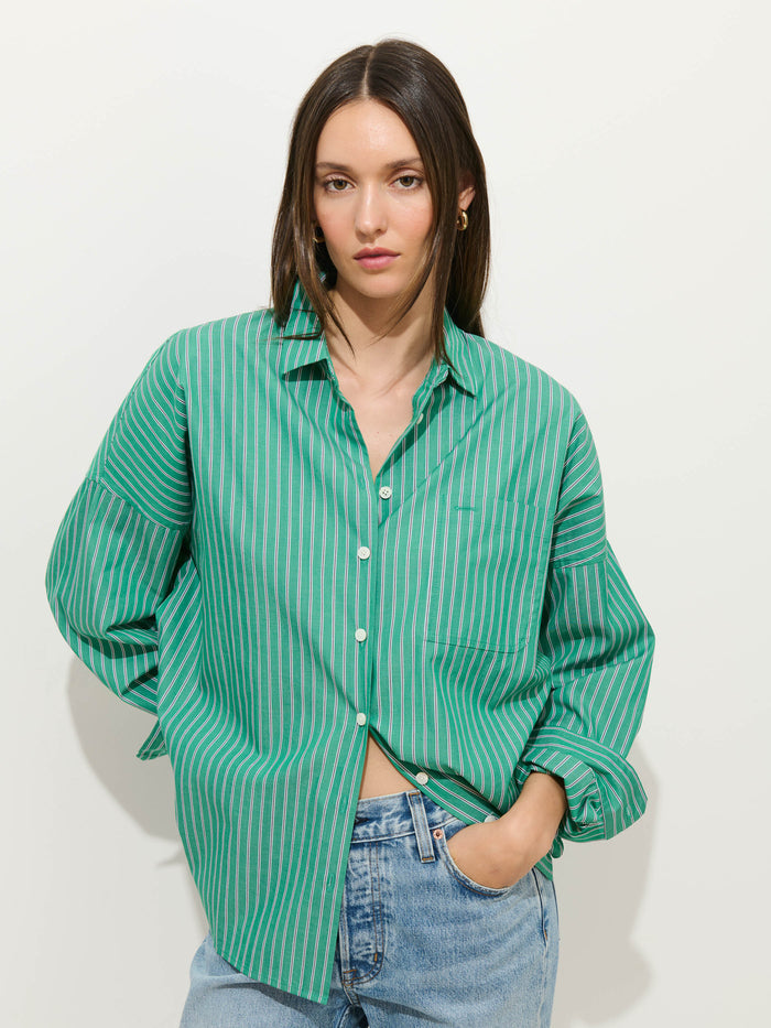 A woman with straight brown hair wears the Jo Striped Shirt In Cotton Poplin—a loose, green and white striped 100% cotton button-up with Troca shell buttons—paired with light blue jeans, standing against a plain white background with one hand in her pocket.