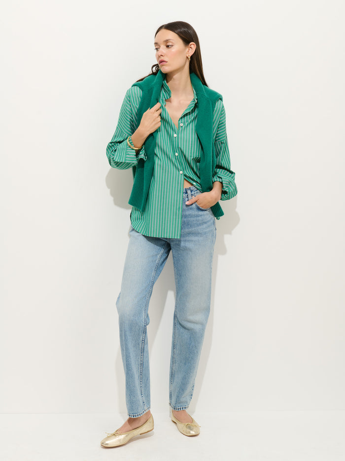 A woman stands against a white wall wearing the Jo Striped Shirt in Cotton Poplin, layered with a green sweater draped over her shoulders, paired with light blue jeans and gold flats. One hand is in her pocket as she looks to the side.