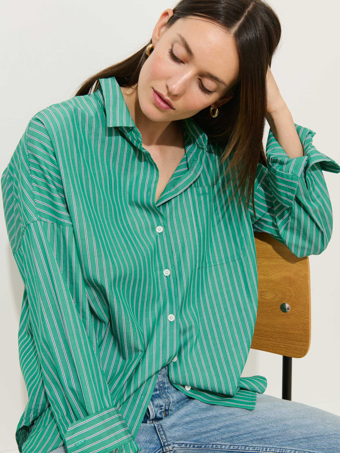 Wearing the Jo Striped Shirt in Cotton Poplin—a relaxed green and white striped button-up crafted from 100% cotton with elegant Troca shell buttons—a woman sits on a wooden chair in jeans, tilting her head down as she gently touches her hair.