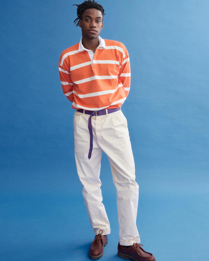 A man stands against a blue background in the J.Press x Alex Mill Rugby Striped Shirt In Cotton, featuring orange and white stripes. He pairs it with cream pants, a purple belt, brown shoes, and looks slightly to the side.