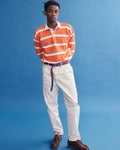A man stands against a blue background in the J.Press x Alex Mill Rugby Striped Shirt In Cotton, featuring orange and white stripes. He pairs it with cream pants, a purple belt, brown shoes, and looks slightly to the side.