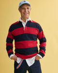 An older man stands smiling with hands in pockets against a yellow background, wearing a J.Press x Alex Mill Rugby Striped Shirt in cotton over a white collared shirt, dark jeans, and a light blue baseball cap.