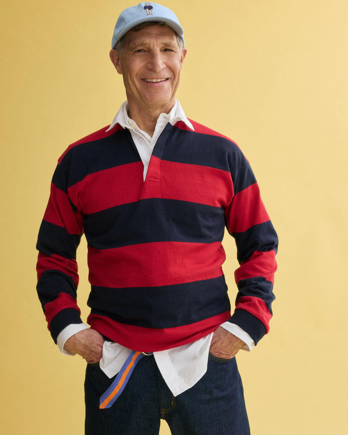 An older man stands smiling with hands in pockets against a yellow background, wearing a J.Press x Alex Mill Rugby Striped Shirt in cotton over a white collared shirt, dark jeans, and a light blue baseball cap.