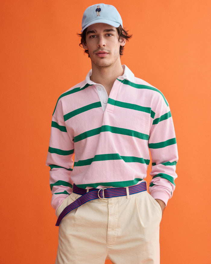 A man in a light blue cap, beige pants, striped belt, and the J.Press x Alex Mill Rugby Striped Shirt in Cotton—pink and green stripes on heavyweight jersey—stands against an orange background.