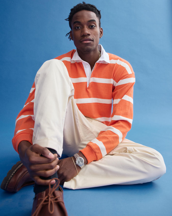 A person sits cross-legged on the floor in a J.Press x Alex Mill Rugby Striped Shirt In Cotton (bright orange and white), cream pants, brown shoes, and a silver watch, set against a plain blue background.