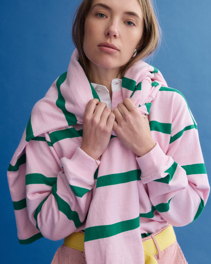 A woman stands against a blue background wearing the J.Press x Alex Mill Rugby Striped Shirt in cotton, paired with a matching sweater over her shoulders and yellow pants. She looks at the camera with a neutral expression.