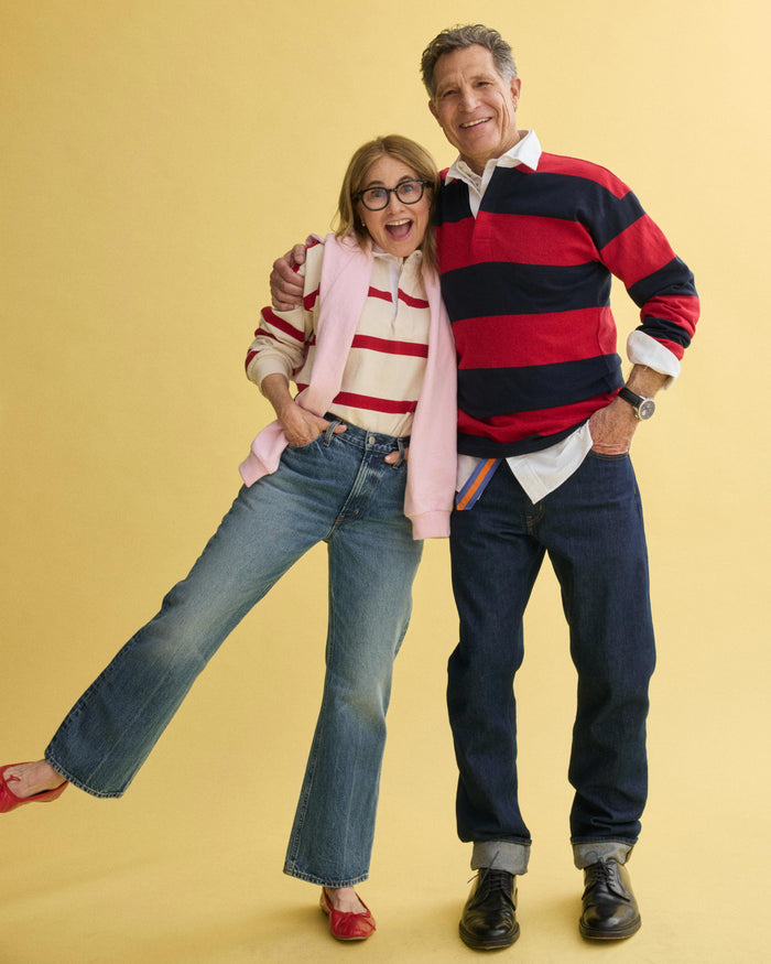 A smiling older couple in matching J.Press x Alex Mill Rugby Striped Shirt In Cotton and jeans stand together against a yellow background. The woman playfully lifts one leg in red shoes while the man has his arm around her shoulders.