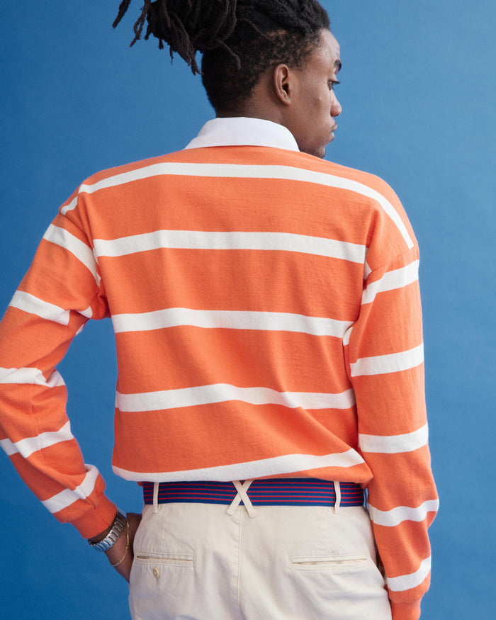 Against a blue background, a person with dreadlocks faces away, wearing the J.Press x Alex Mill Rugby Striped Shirt in Cotton (orange and white stripes), cream pants, a blue and orange striped belt, and a watch.