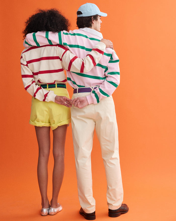 Two people stand arm in arm, facing away from the camera, wearing J.Press x Alex Mill Rugby Striped Shirts in cotton and light pants or shorts. Their pinkies are linked behind their backs, set against an orange background.