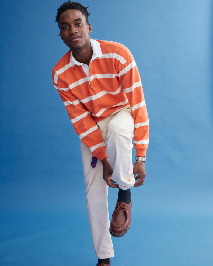 A man in a J.Press x Alex Mill Rugby Striped Shirt In Cotton bends to adjust his black sock. He wears it with cream pants and brown shoes, standing against a plain blue background with a slight smile.