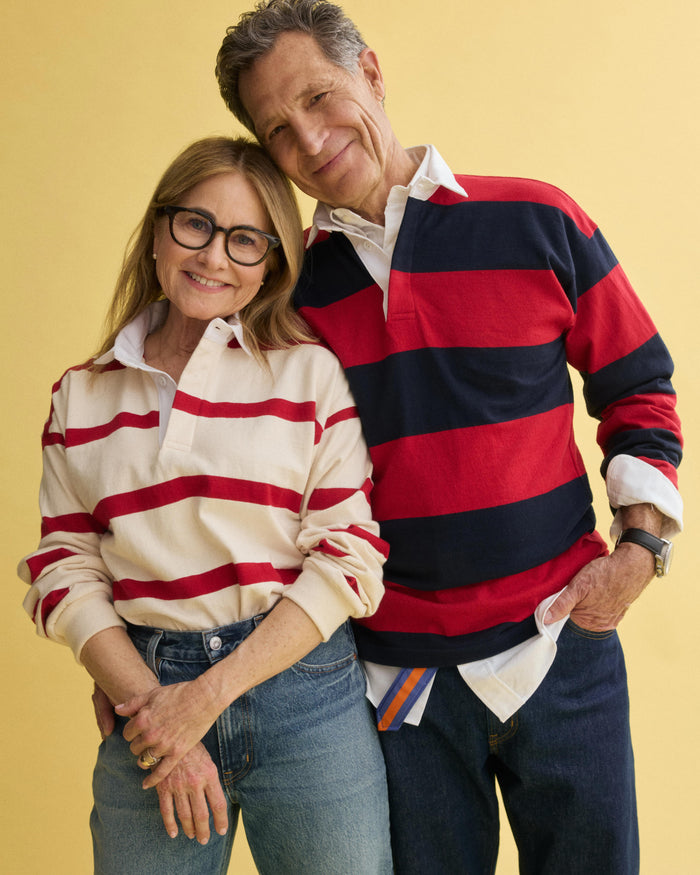 A smiling older couple poses against a yellow background, both in J.Press x Alex Mill Rugby Striped Shirt In Cotton and jeans. The woman, wearing glasses, leans against the man’s side as he wraps his arm around her shoulders.