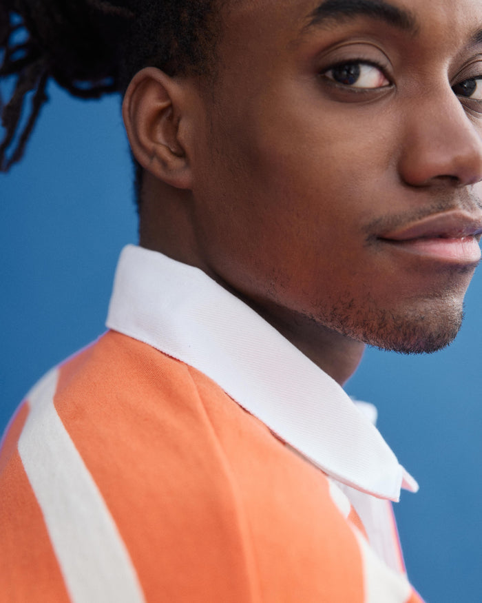 A close-up of a person with brown skin and short facial hair wearing the J.Press x Alex Mill Rugby Striped Shirt In Cotton, featuring orange and white stripes, looking over their shoulder against a blue background.