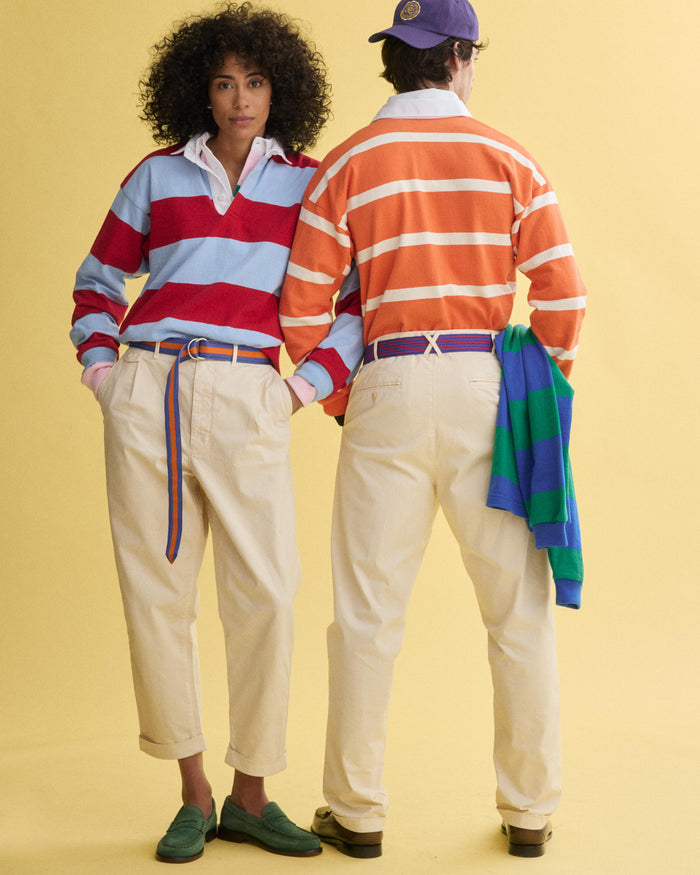 Two people in cream pants and the J.Press x Alex Mill Rugby Striped Shirt in Cotton—a red and blue version worn facing forward, an orange and white one with a green sweater tied at the waist, turned away—stand against a yellow background.