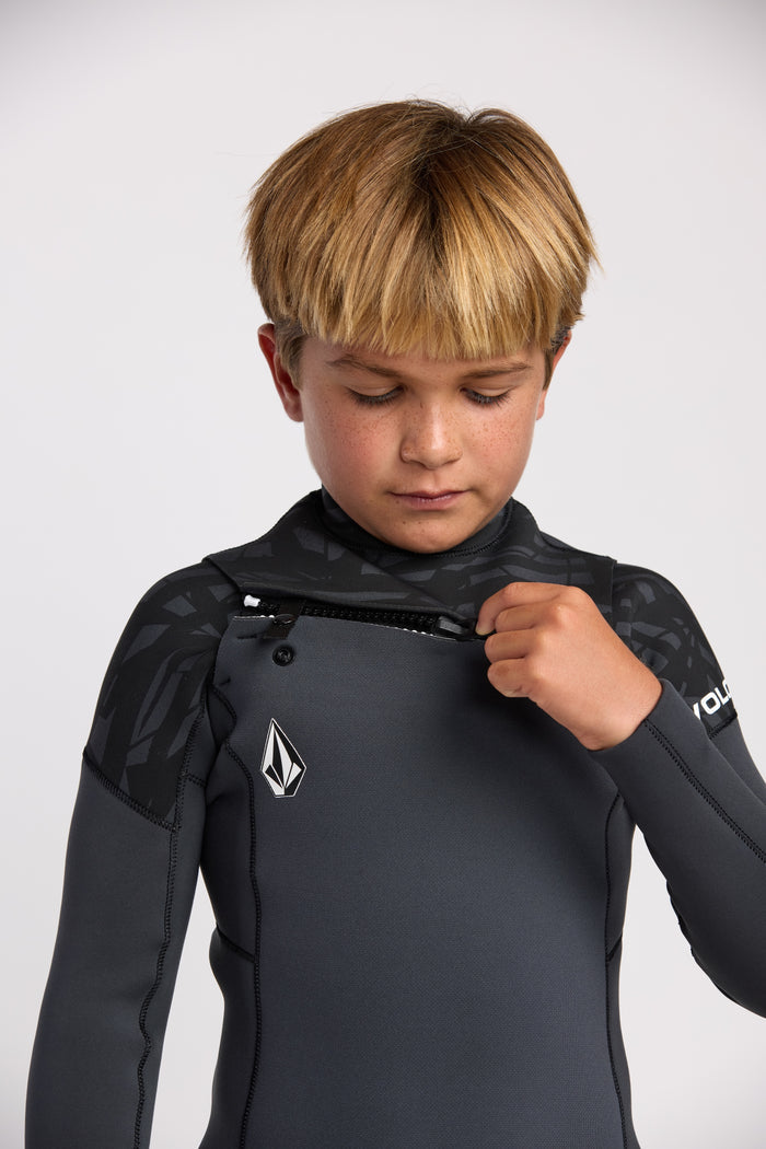 A blonde boy stands against a plain light background, pulling up the front zipper of the Volcom Kids' 3/2 Modulator Chest Zip Full Wetsuit, ready for his next surf adventure.