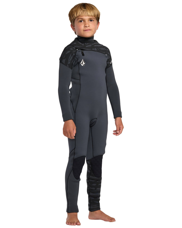 A young boy with short blond hair stands barefoot in a Volcom Kids' 3/2 Modulator Chest Zip Full Wetsuit, featuring geometric patterns on the shoulders and knees. Facing forward, he’s ready to surf with his arms relaxed by his sides.