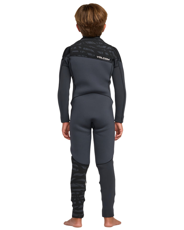 A child stands barefoot, facing away, wearing the Volcom Kids' 4/3 Modulator Chest Zip Full Wetsuit in black and dark gray with a textured pattern on the shoulders and lower left leg. The Volcom logo appears on the upper back.