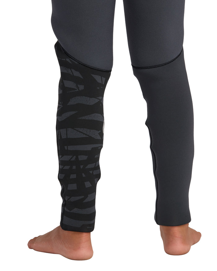 A child stands barefoot, seen from the back, wearing Volcom's Kids' 4/3 Modulator Chest Zip Full Wetsuit featuring fitted dark legs and a black and gray geometric pattern on one leg.