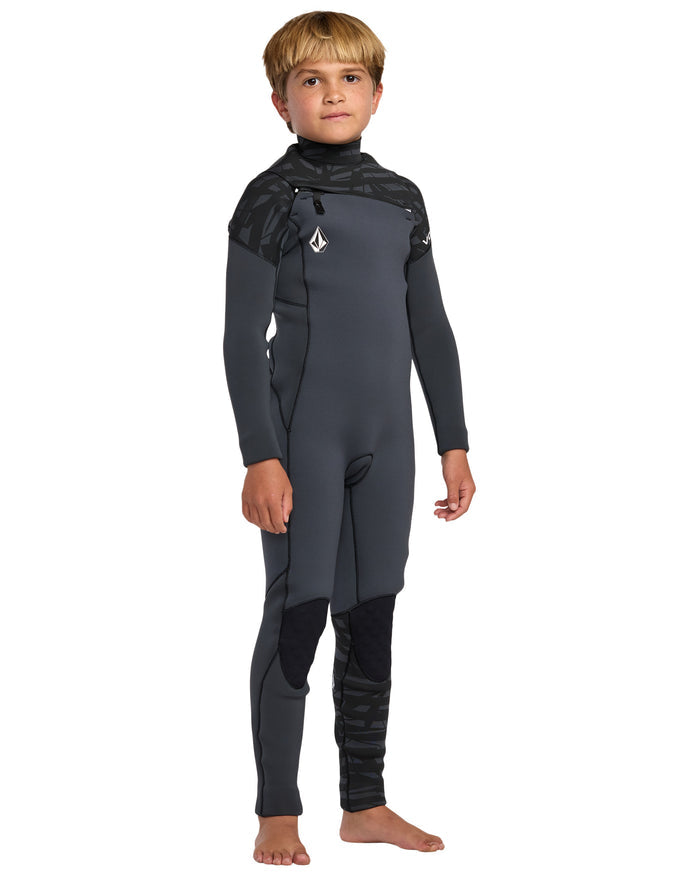 A young boy stands barefoot in the Volcom Kids' 4/3 Modulator Chest Zip Full Wetsuit with dark gray fabric, black accents, and a chest logo, posing confidently against a white background.