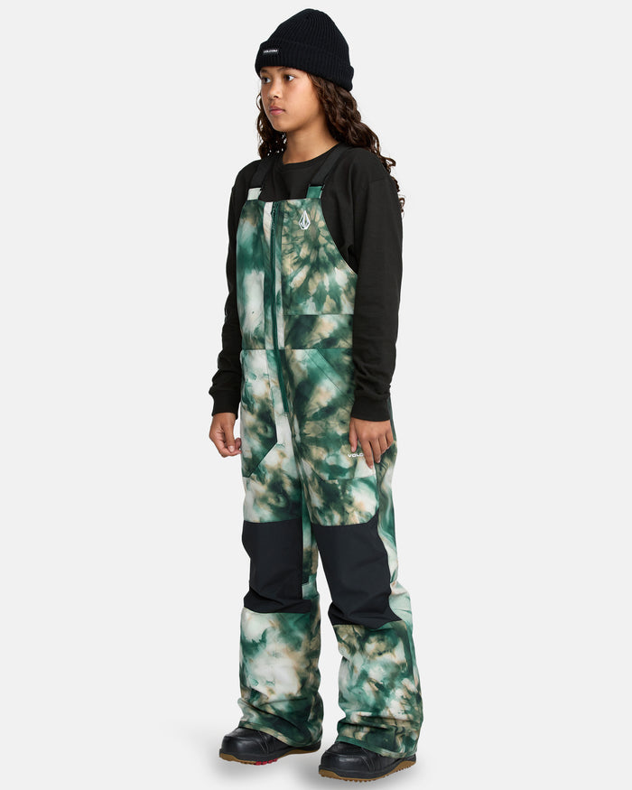 A child wearing a black beanie, black long-sleeve shirt, and the Kids' Barkley Insulated Bib Overall in green tie-dye with black knee patches stands and looks slightly to the side.