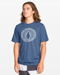 A young man with long brown hair wears a Volcom Men's Men blue crew neck short sleeve tee featuring a white circular logo on the chest and black-and-white shorts, standing against a plain white background.