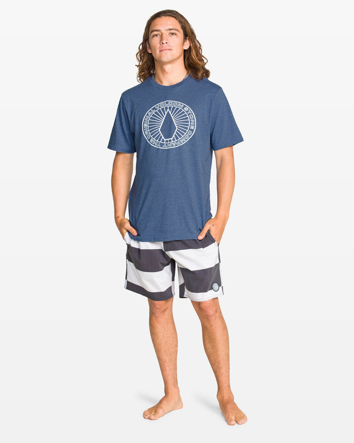 A young man with long brown hair stands barefoot against a plain background, wearing the Volcom Men's Men crew neck blue tee and black-and-white striped board shorts. Hands in his pockets, he faces forward with a relaxed look.