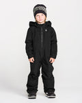 A child models the Volcom Kids’ Little Little Flurry One Piece Snow Suit in black, paired with a black and white knit hat and dark shoes, standing facing forward against a plain white background.