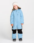 A child wearing the Volcom Kids’ Kid’s Little Flurry One Piece snowsuit—shown here in light blue and black with 10K/10K protection and recycled V-Science fabric—stands facing forward against a plain white background, accessorized with a white beanie and dark boots.