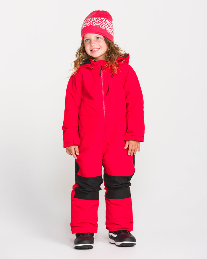 A young child stands smiling against a plain white background, wearing the Volcom Kids’ Kid’s Little Flurry One Piece snowsuit, a red hat with white writing, black gloves, and winter boots.