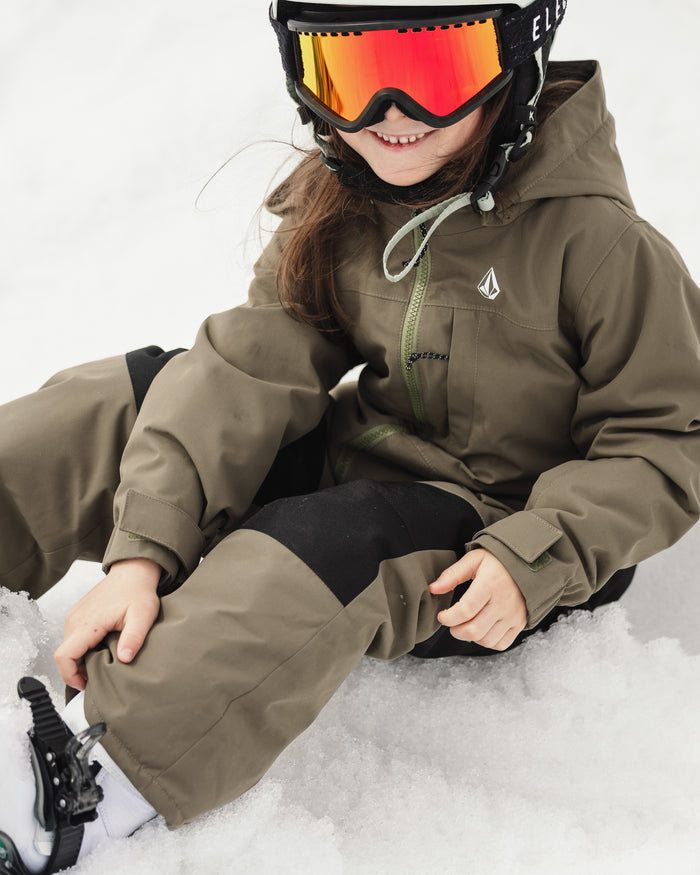 A child sits on the snow, smiling as they adjust their boot while bundled up in the Volcom Kids’ Little Flurry One Piece—a cozy kids’ snow suit made from recycled V-Science materials. The child is also wearing an olive green ski jacket, black snow pants, and a helmet with large reflective orange ski goggles.
