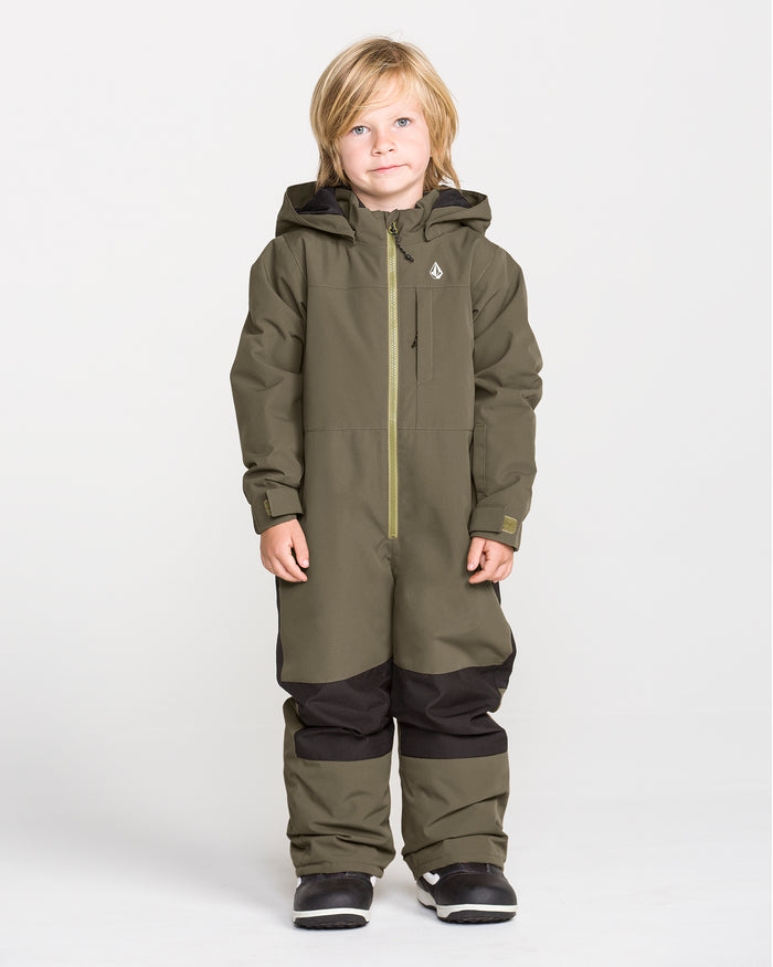 A young child with blonde hair stands against a plain white background, dressed in the Volcom Kids’ Little Flurry One Piece snowboard suit in olive green, which features black knee patches and black boots. This insulated snowsuit is crafted from recycled V-Science materials for warmth and sustainability.