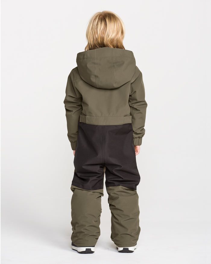A child with long blonde hair stands facing away, wearing the Volcom Kids’ Kid’s Little Flurry One Piece snowboard suit in olive green and black, crafted from recycled V-Science materials, paired with white shoes against a plain white background.