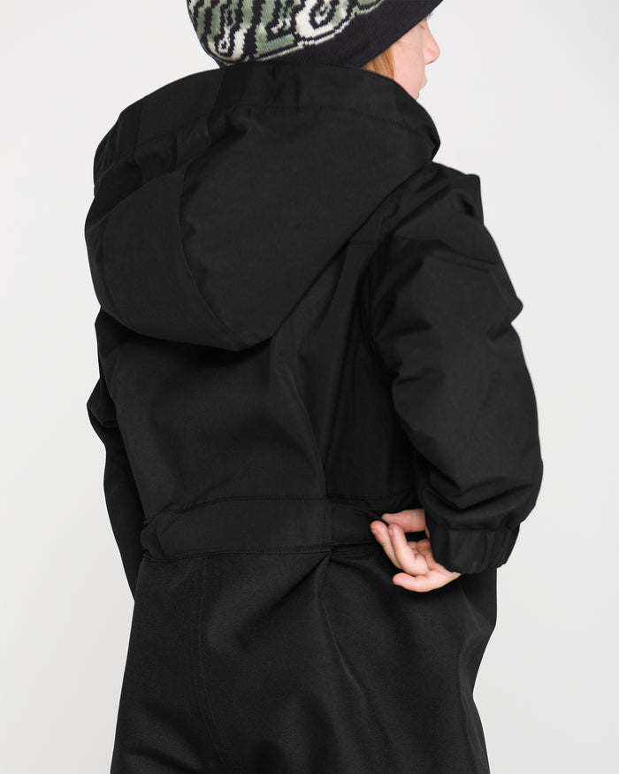 A child faces away against a plain light background, wearing the Volcom Kids’ Little Little Flurry One Piece Snow Suit in black with a patterned winter hat, holding the back of the suit with one hand.