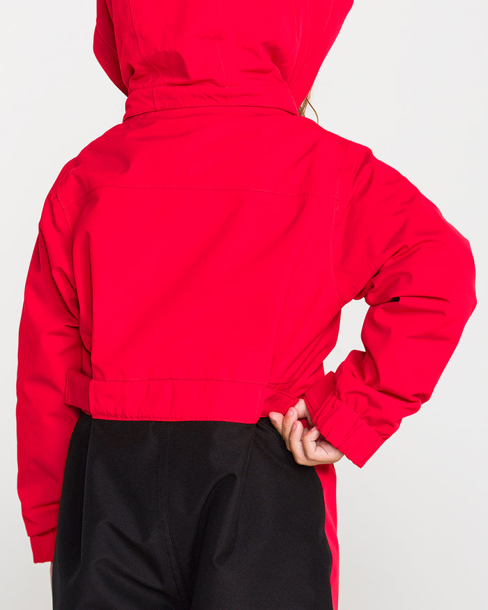 A child stands with their back to the camera, one hand on their hip, wearing the Volcom Kids’ Little Flurry One Piece in bright red and black pants against a plain light background, showcasing an insulated design crafted from recycled V-Science fabric for enhanced 10K/10K protection.