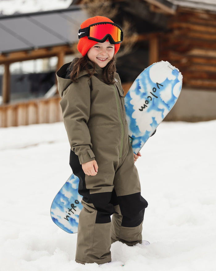 A smiling child stands on the snow in a green Volcom Kids’ Little Flurry One Piece made from recycled materials, paired with a red beanie and ski goggles, holding a blue and white snowboard in front of a wooden building.