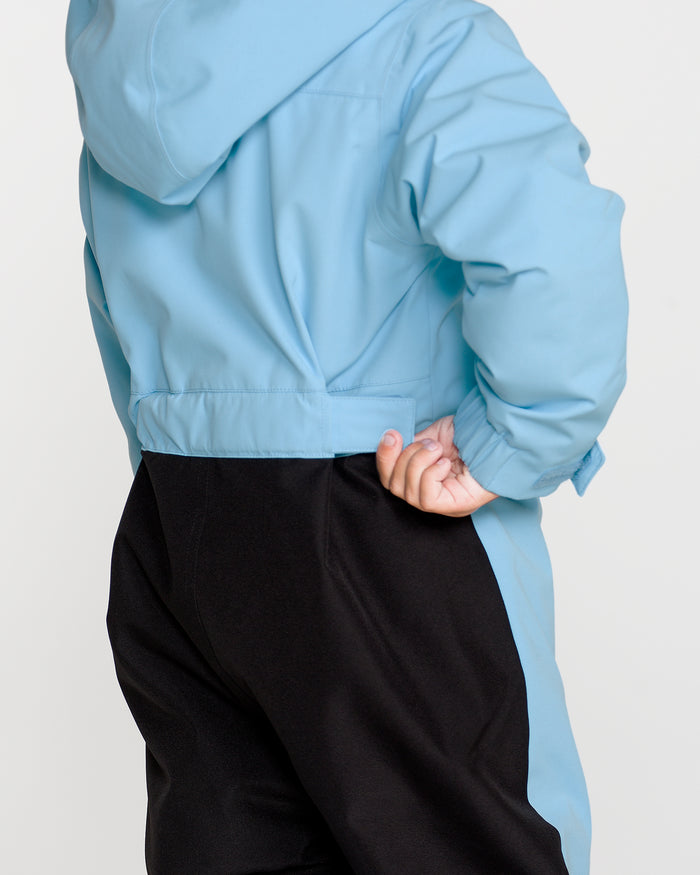 A child wearing the Volcom Kids’ Kid’s Little Flurry One Piece in light blue—crafted from recycled V-Science fabric—is pictured from behind, standing with one hand on their hip.