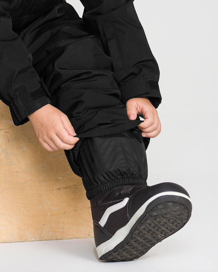 A child wearing the Volcom Kids’ Little Little Flurry One Piece Snow Suit sits on a wooden block while adjusting the elastic cuff of their pant leg.