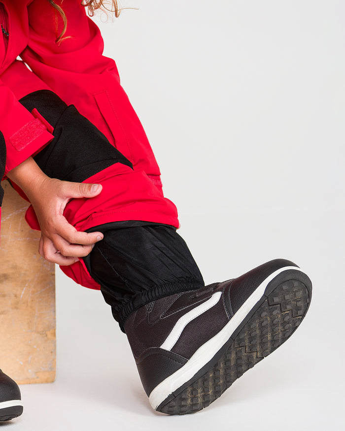 A child in a red jacket adjusts the cuff of their pant leg, revealing black snow boots with white details. Only the lower half of the body is visible, showcasing the Volcom Kids’ Little Flurry One Piece, designed for 10K/10K protection.