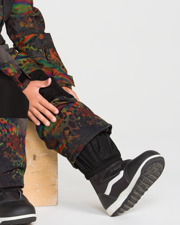 A child wearing the Volcom Kids’ Little Flurry One Piece with colorful patterns sits on a wooden block, black sneakers on their feet and hands resting on their leg, against a plain white background.