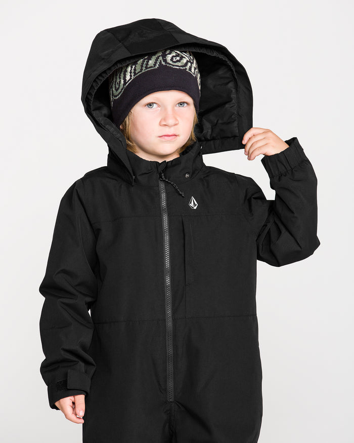 A young child wearing the Volcom Kids’ Little Little Flurry One Piece Snow Suit and a patterned beanie looks at the camera, holding the hood with one hand against a plain light background.