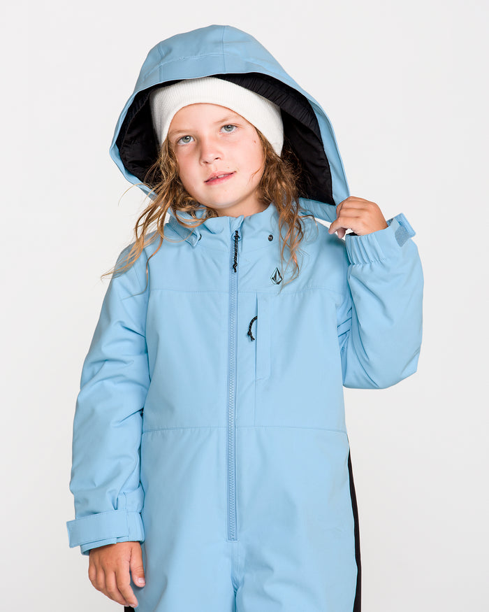 A young child with long hair wears a white beanie and the Volcom Kids’ Little Flurry One Piece—a light blue hooded snowsuit made from recycled V-Science material—looking slightly upward while holding the side of the hood. The background is plain white.