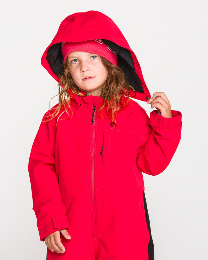 A young child with long hair wears the Volcom Kids’ Little Flurry One Piece in bright red, along with a matching hat, holding the hood with one hand while standing against a plain white background. This insulated snowsuit boasts 10K/10K protection and is crafted from recycled V-Science fabric for excellent warmth and sustainability.