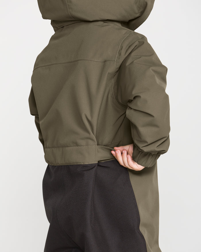 A child wearing the Volcom Kids’ Little Flurry One Piece snowboard suit in olive green, crafted from recycled V-Science fabric, stands with their back to the camera and holds the waistband of their jacket with one hand.