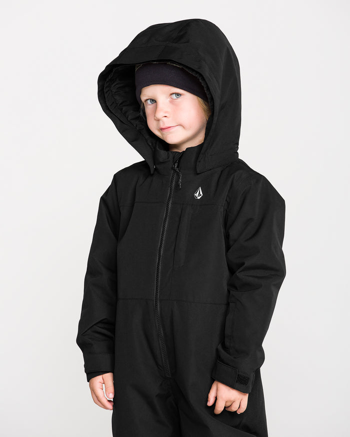 A young child wearing the Volcom Kids’ Little Flurry One Piece Snow Suit and a black beanie stands against a plain white background, looking slightly to the side with a neutral expression.