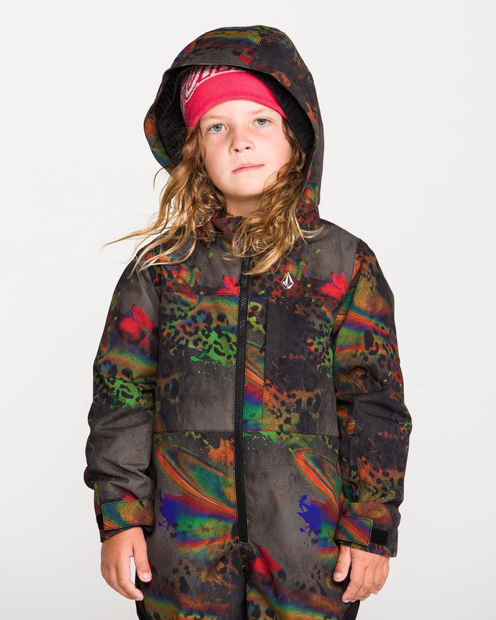 A young child with long hair stands against a plain white background, wearing the Volcom Kids’ Kid’s Little Flurry One Piece—a colorful, patterned hooded one-piece crafted from recycled V-Science materials for warm insulation—paired with a red beanie.