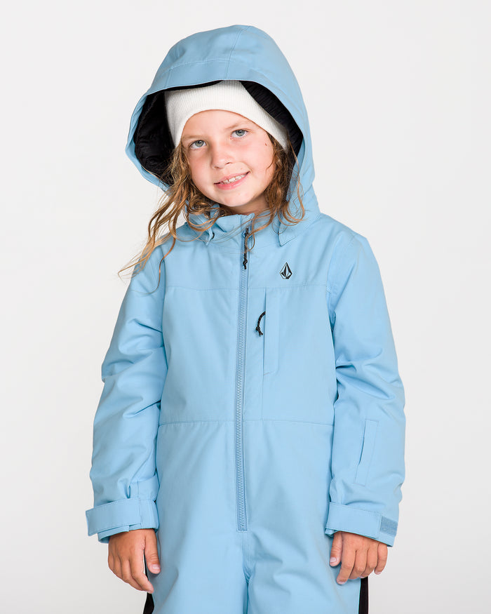 A young child smiles at the camera against a plain white background, dressed in the Volcom Kids’ Little Flurry One Piece—a light blue hooded winter suit featuring recycled V-Science fabric and 10K/10K protection—paired with a white beanie.